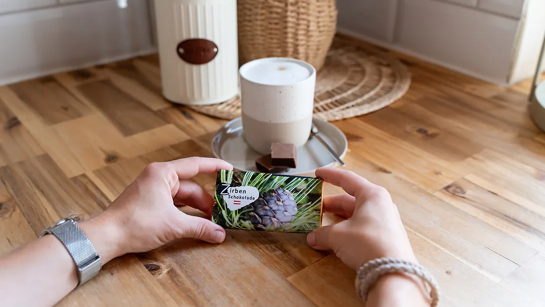 Person hält eine Packung Zirbenschokolade über einem Holztisch, neben einer Tasse auf einem Untersetzer.